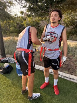 Two players from the OKC Bulldogs flag football team adjust their uniforms, with the player on the right prominently displaying the "Kendu Bulldogs" logo on his jersey, representing the team's official sponsorship by the KenduInu community as mentioned in the post.