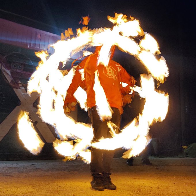A performer in an orange jacket skillfully spins flaming props, creating a bright, circular trail of fire in a nighttime performance.