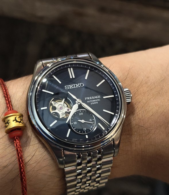 A close-up of a wrist wearing a Seiko Presage Automatic 3 Days watch with a black open-heart dial showing the movement, paired with a red string bracelet with a gold bead.