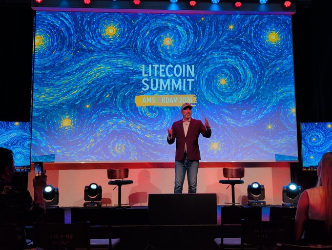 The image shows a man standing on stage, giving a speech or presentation while wearing a suit and tie. There are several people sitting in chairs around him, possibly listening to his talk or waiting for their turn to speak. The stage has a large screen behind the speaker that displays a starry night sky with swirls of light, creating an immersive atmosphere for the event. The image also includes text that reads "Litecoin Summit" and "Ams.ROOM.ROAM.2020", indicating the name of the event or gathering taking place.