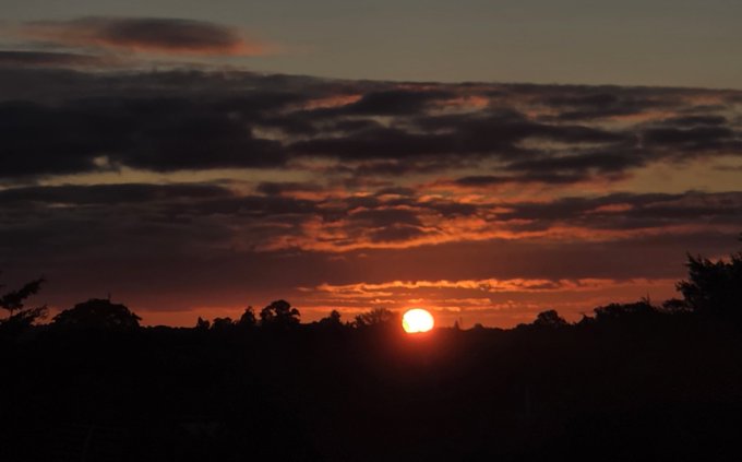 A vibrant sunrise glows over a silhouetted treeline, with the bright orange sun resting just above the horizon amid scattered clouds tinted in fiery hues. The image perfectly complements the post's message of fresh beginnings and a hopeful new week, visually echoing the optimism of welcoming the day with the XRP community.