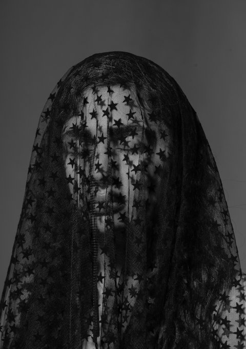 A black-and-white artistic portrait of a person whose face is partially obscured by a sheer black veil patterned with stars, set against a gray background.