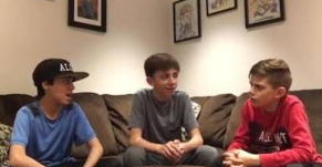 Three young men sitting on a couch having a conversation, with one wearing a black cap and blue shirt, one in a gray shirt, and one in a red shirt, with framed pictures visible on the wall behind them.