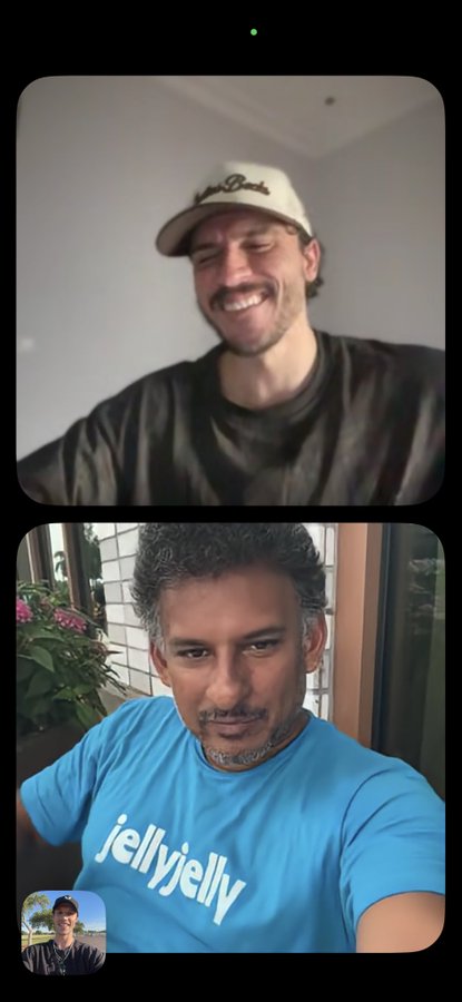 A video call screenshot showing two men: the top man wears a 'Peach Beach' cap and smiles with eyes shut, while the bottom man wears a blue 'jellyjelly' t-shirt and smiles at the camera.