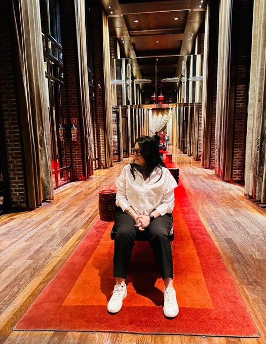 A woman in a white shirt and black pants sits on a bench in an ornate hallway with wooden floors and a red runner rug.