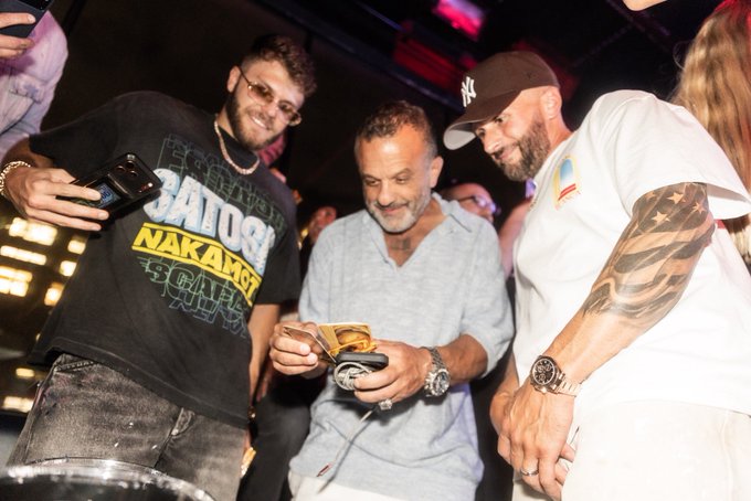 Three men at what appears to be a nightclub or social event; the center man in a gray shirt is smiling while looking at a phone, flanked by a man in a black 'SATOSHI NAKAMOTO' t-shirt and sunglasses on the left, and a bearded man in a white NY cap and white t-shirt with a tattooed arm on the right.