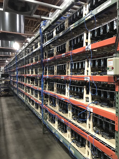 A photograph of a large-scale industrial mining facility showing rows of mining rigs mounted on orange shelving units, connected by extensive blue cabling and ductwork in a warehouse setting.