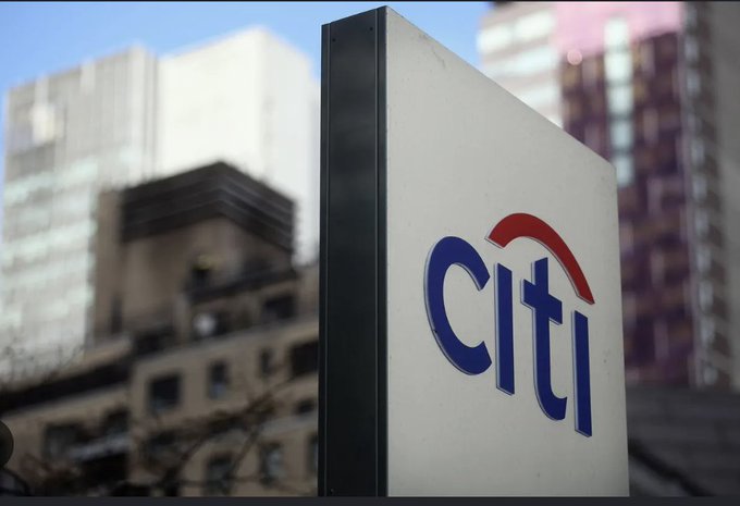 A close-up photograph of a Citi (Citibank) sign mounted on a building, with the blue 'citi' logo and red arch visible against a blurred urban background of skyscrapers.