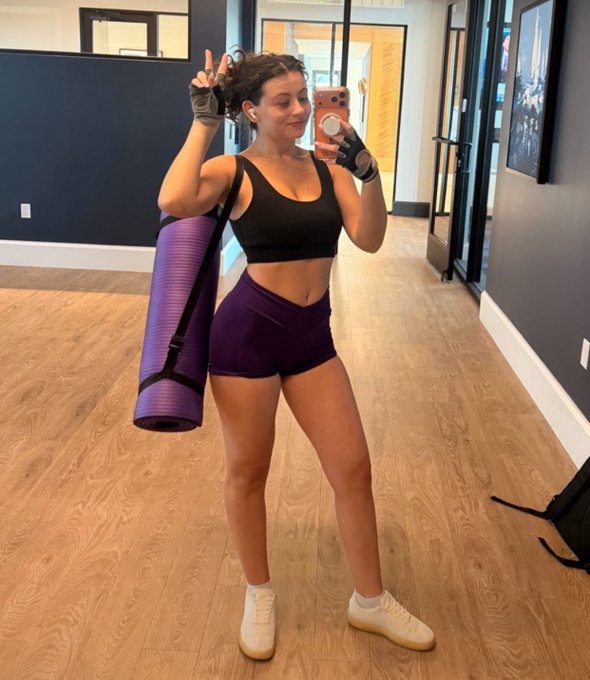 A woman poses in a gym mirror selfie wearing a black sports bra, purple shorts, and fingerless gloves, carrying a rolled-up purple yoga mat while flashing a peace sign. The image aligns with the post's caption, visually reinforcing the gym workout aspect of her Saturday routine, though no Bitcoin or crypto-related text or imagery is visible in the photo itself.