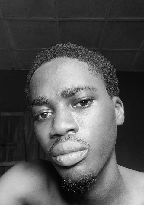 Black and white close-up selfie of a young man with short hair, looking directly at the camera with a neutral expression in a dimly lit room.