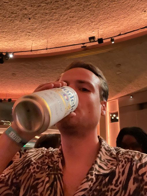A young man in a patterned shirt takes a selfie while drinking from a large beverage cup at what appears to be a venue or event.