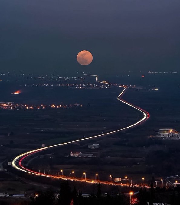 A nighttime landscape photograph showing a full moon over a winding road with streaks of car lights illuminating the path through a dark valley.