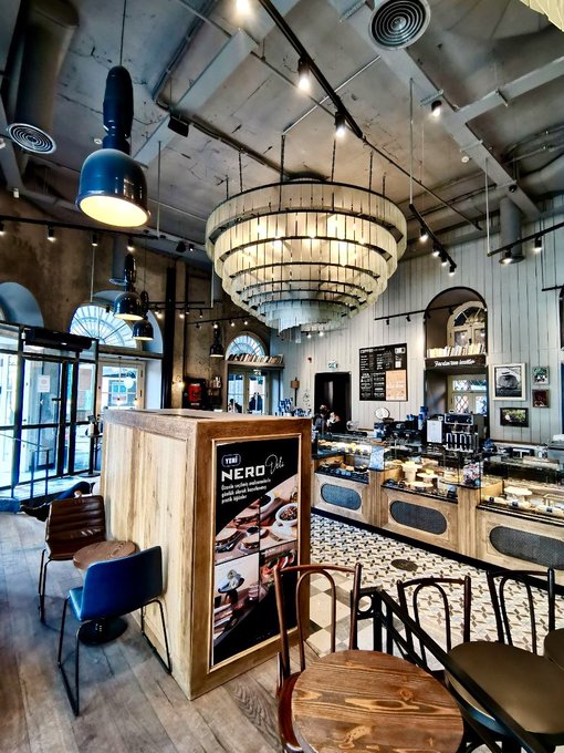 The interior of 'THE NERO' café and bakery showcases a large tiered chandelier, industrial ceiling, patterned tile flooring, and a wooden display counter filled with pastries alongside a branded sign.