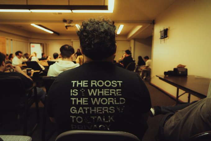 The back of a person wearing a black t-shirt that reads 'THE ROOST IS WHERE THE WORLD GATHERS TO TALK PRIVACY' at a crowded indoor event.
