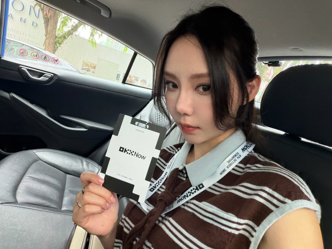 A young woman sitting in the backseat of a car holding up an 'OKX Now' promotional card, wearing a brown and white striped shirt with a lanyard.