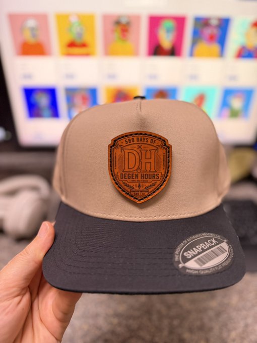 The image features a person holding a baseball cap that has a wooden shield patch on it. The cap appears to be made of black, white, and tan colors. There are several other hats displayed around the main hat being held by the person. These hats can be seen in various positions and orientations, showcasing their different designs and styles.