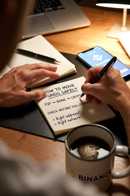 A person's hands write 'HOW TO MOVE FUNDS SAFELY P2P → BANK → CRYPTO' in a notebook on a desk, accompanied by a smartphone showing the Binance app and a Binance-branded coffee mug.