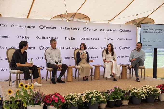 A panel of five people sits on a stage at an event branded 'Out East Summit' and 'Canton'. The backdrop also displays 'The Tie', and a screen reads 'Should We Be Optimistic' about institutions.