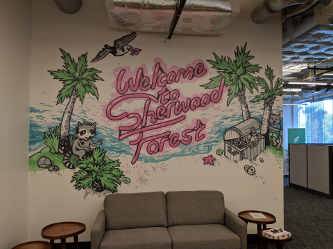 A mural on an office wall reading 'Welcome to Sherwood Forest' in pink script, surrounded by illustrations of palm trees, a raccoon, a treasure chest, and a bird, located above a brown sofa.