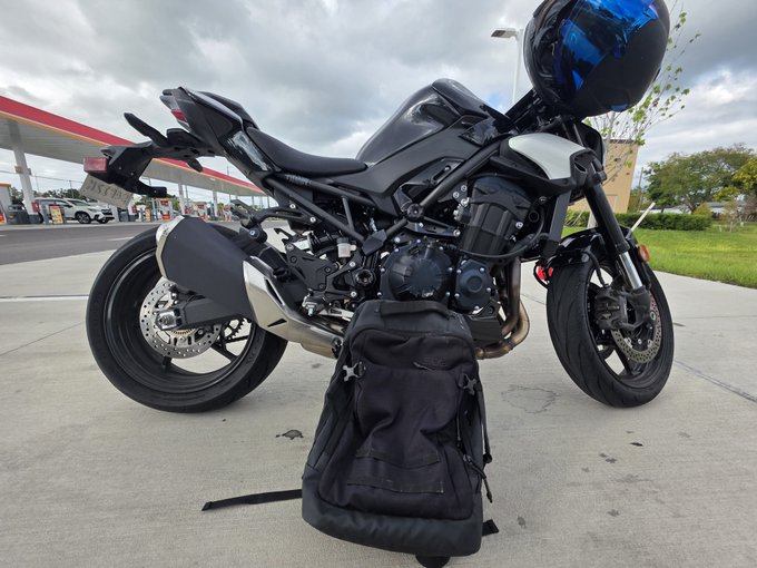 A black motorcycle is parked at a gas station with a black backpack resting on the ground in front of it, and a black and blue helmet placed on top of the seat.