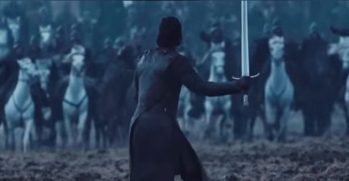 A man in a dark coat stands with his back to the camera, holding a sword, facing a large group of figures on horseback in a foggy, dark setting.