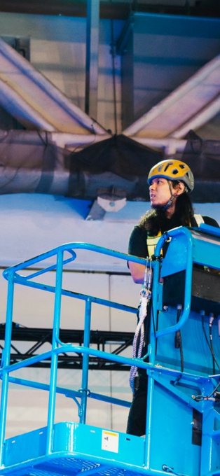 A woman wearing a yellow safety helmet and a black shirt stands on a blue elevated work platform, looking upward under a ceiling with skylights, appearing to be at an outdoor work site.