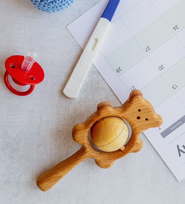 A flat-lay photograph displays a wooden teddy bear rattle toy alongside a red pacifier, a pregnancy test stick, and a calendar on a light surface.