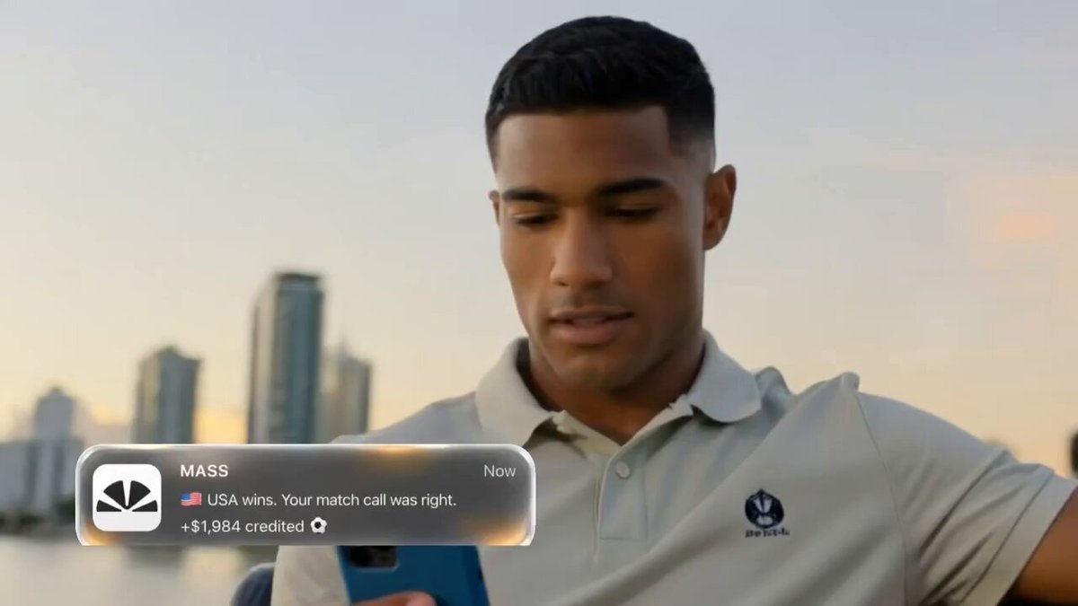 The image shows a man sitting down, looking at his cell phone screen while wearing a white polo shirt. The background of the image features a cityscape with tall buildings, suggesting that he might be in an urban setting or near a waterfront. The text on the image reads "If you were already aware, @Polymarket is now live on @massdotmoney". This implies that the man has recently become interested in using the mass dot money app and is likely exploring its features or checking for updates.