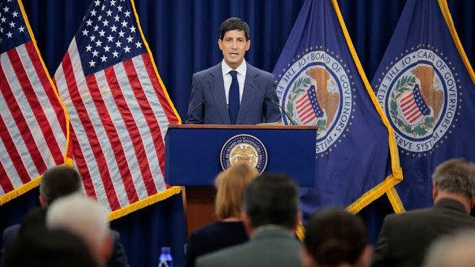 A man in a suit stands at a podium bearing the Federal Reserve seal, delivering remarks against a backdrop of American flags and Federal Reserve banners, as an audience listens. The image relates to the post's caption by depicting a Federal Reserve official whose hawkish or dovish stance can significantly influence crypto market sentiment.