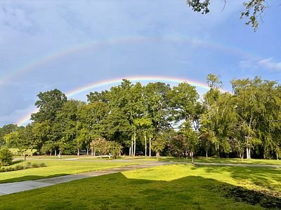 A scenic landscape showing a vibrant rainbow arching across a cloudy sky over lush green trees and a grassy field with a paved path.