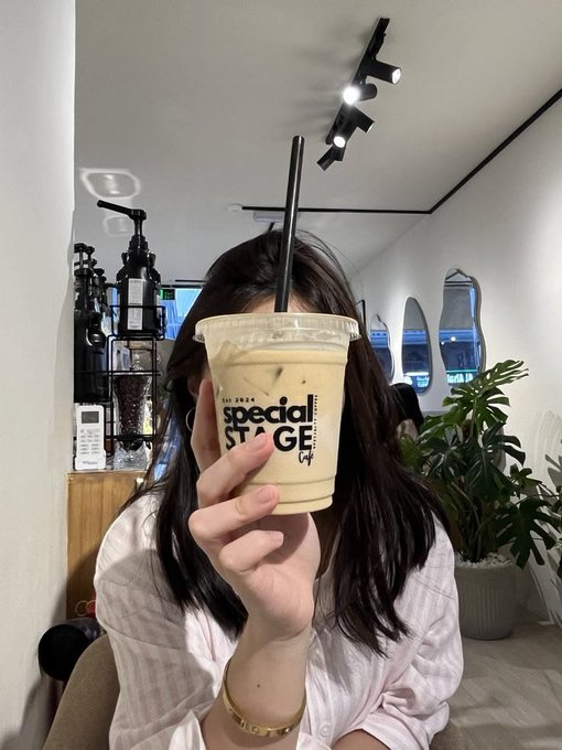 A woman holds a cup of iced coffee from 'Special Stage' café in front of her face, wearing a white shirt.