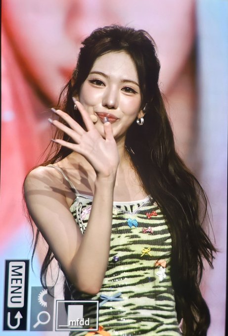 A young woman with long dark hair smiles and waves her hand while wearing a striped dress; the image has UI overlays including "MENU" and "mfdd".