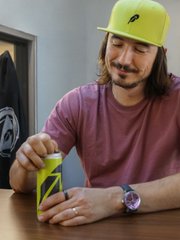 A man wearing a neon yellow cap and maroon shirt holds a green and black Monster energy drink can at a table.