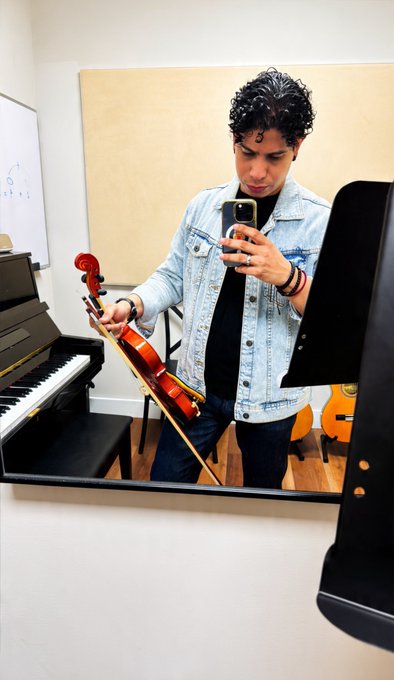 A mirror selfie of a young man with curly hair wearing a light blue denim jacket over a black shirt, holding a violin and bow in one hand and a smartphone in the other.