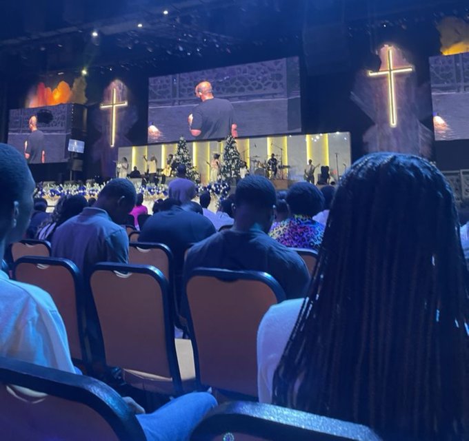 A photograph from the audience perspective inside a large church or auditorium, showing people seated in chairs watching a man speaking on stage with illuminated cross decorations in the background.