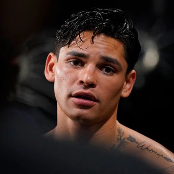 A close-up portrait of a young man with dark wet hair, glistening skin, and a tattoo visible on his shoulder/chest area against a dark background.