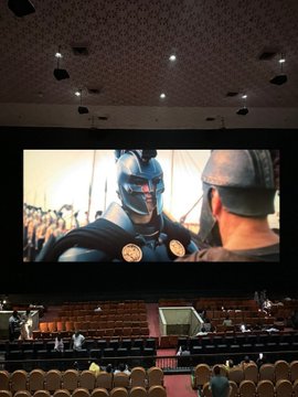 A person is likely taking a photo of a Batman movie projected on a large cinema screen, with the theater's seating visible below and a few audience members scattered around. The "CEX:" and "UEX:" captions appear unrelated to the Batman imagery and instead seem to reference exchange-related listings or comparisons.