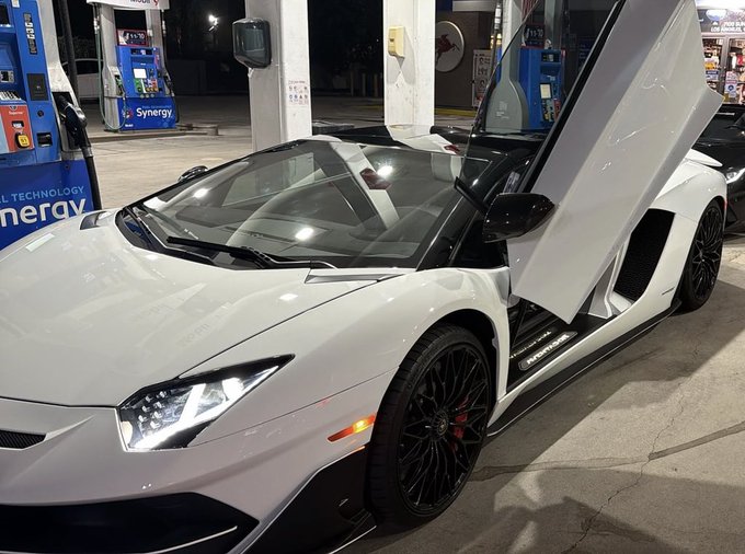 A white Lamborghini Aventador with its scissor doors open parked at a Synergy gas station at night.