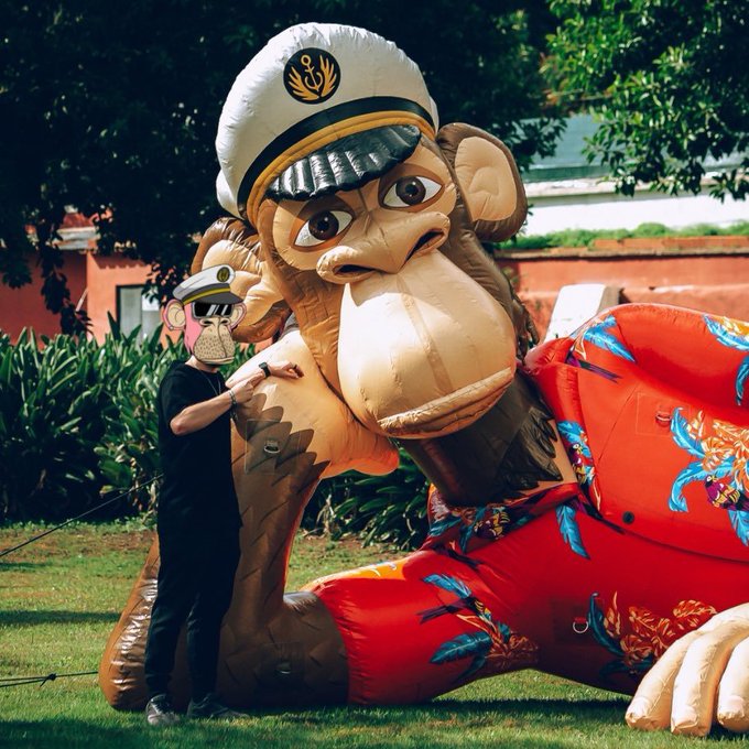 A person stands next to a massive, inflatable monkey character dressed in a captain's hat and a red floral shirt. The image humorously visualizes the concept of "touching grass" by showing someone interacting with an oversized, whimsical object in a grassy outdoor setting.