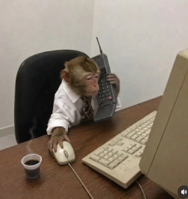 A small monkey wearing a white dress shirt and tie sits at a desk using a vintage computer mouse and holding a large retro mobile phone to its ear, with a small cup of coffee beside it.