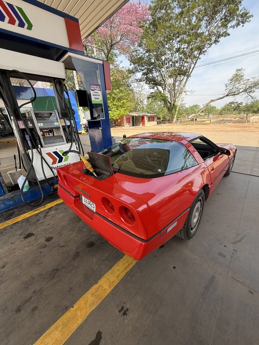 A red Chevrolet Corvette sports car parked at a gas station, with a laptop resting on its rear bumper near the fuel cap.