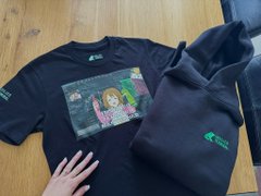 Two black garments—a t-shirt featuring colorful cartoon characters and a hoodie—both displaying a small green logo, rest on a wooden surface. The matching apparel pairs nicely with the casual "GM" (Good Morning) greeting.