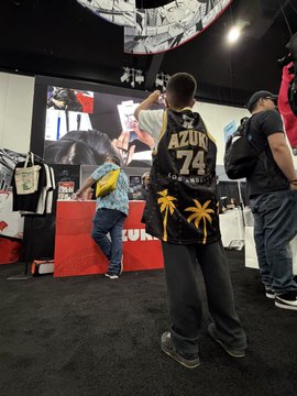 A young boy poses in front of a screen at the Azuki booth, wearing an oversized black jersey with "AZUKI 74" printed on the back and a palm tree design on the front, while browsing the exhibit at Anime Expo. The image captures the welcoming, stress-free atmosphere that made Azuki's booth stand out among the convention's many exhibits.