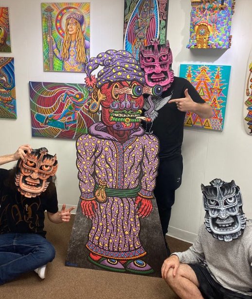 Two people posing in an art gallery, one seated wearing a traditional mask and holding a drink, the other in a black shirt wearing a pink mask, standing next to a large colorful cutout figure with tribal-style designs, surrounded by vibrant artwork on the walls.