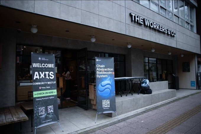 The exterior of a venue called 'THE WORKERS & CO' in Tokyo, Japan, featuring banners for an 'AXIS' community meetup and a presentation on the 'Chain Abstraction Stablecoin System' with a QR code.