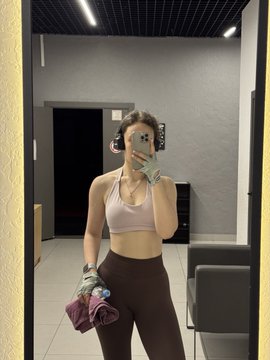A mirror selfie of a woman in athletic wear (pink sports bra and brown leggings) holding a phone with a peace sign, standing in what looks like a gym or apartment lobby.