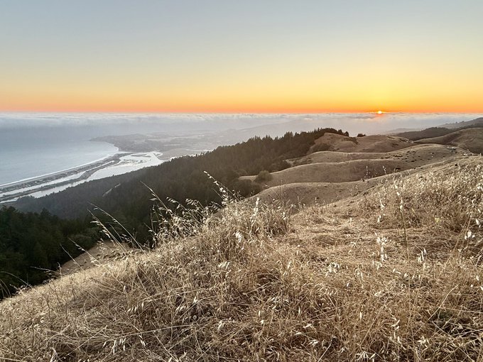 Golden sunset blankets rolling coastal hills and a distant ocean as the sun dips below the horizon, casting a serene glow over the landscape. The tranquil scene captures the stillness and reflection fitting for a peaceful Sabbath.