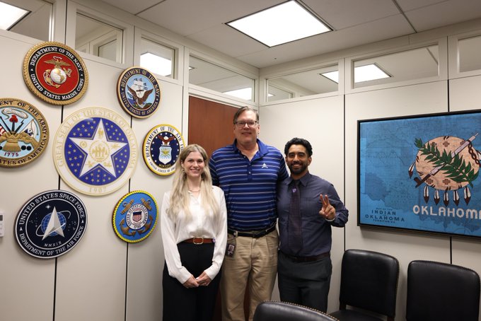 Three people pose in front of a wall displaying various U.S. military and state department seals next to a screen showing an Oklahoma state emblem.