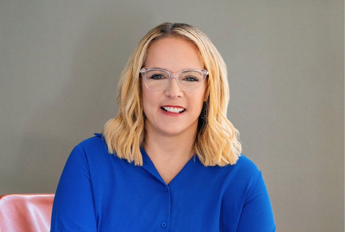 A professional headshot of a smiling blonde woman with glasses wearing a blue button-up shirt against a gray background.