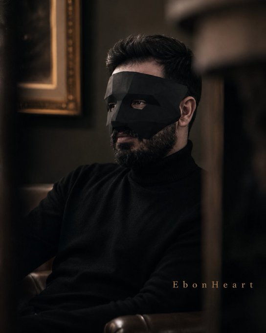 A bearded man wearing a black geometric mask and a black turtleneck sits in a dimly lit room with a framed painting behind him; the text 'EbonHeart' appears in the lower right.